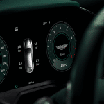 A digital car dashboard display shows 0 mph speed, tire pressures, a central car graphic, “Sport” mode with the Aston Martin logo, Carplay Ultra integration, a temperature of 23°C, and various dashboard indicators.