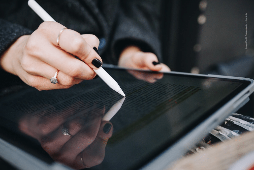 A person with dark nail polish uses a stylus and the Scribble feature on an iPad to write or highlight text on the tablet screen, which reflects their hand. They wear rings and a dark sleeve.