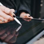 A person with dark nail polish uses a stylus and the Scribble feature on an iPad to write or highlight text on the tablet screen, which reflects their hand. They wear rings and a dark sleeve.