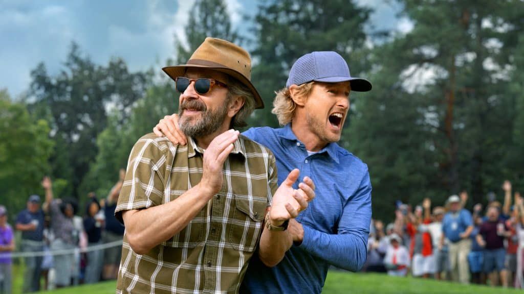 Two men stand outdoors on a golf course—one in a brown hat and sunglasses smiling, while the other in a blue shirt and cap shouts excitedly—capturing the thrill of an Apple TV+ golfing event with a cheering crowd and trees in the background.