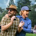 Two men stand outdoors on a golf course—one in a brown hat and sunglasses smiling, while the other in a blue shirt and cap shouts excitedly—capturing the thrill of an Apple TV+ golfing event with a cheering crowd and trees in the background.