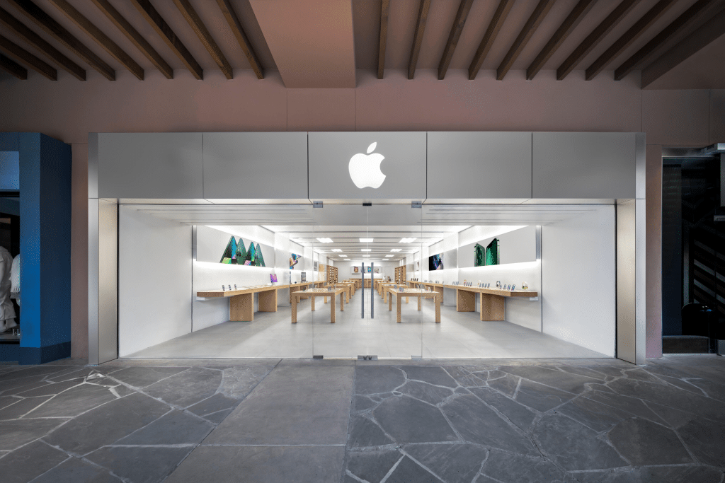 A modern Apple Store at La Encantada with a large illuminated Apple logo above the entrance, glass doors, and an interior featuring wooden tables displaying products in a bright, minimalist space.