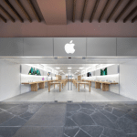 A modern Apple Store at La Encantada with a large illuminated Apple logo above the entrance, glass doors, and an interior featuring wooden tables displaying products in a bright, minimalist space.