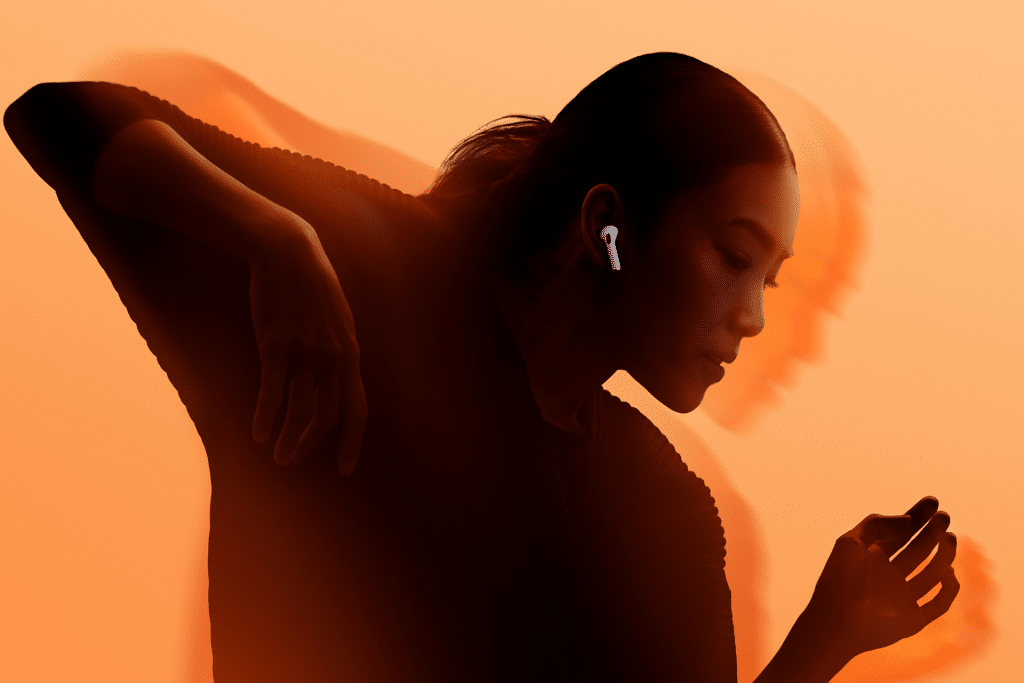 A person with long hair in a ponytail wears AirPods, showcasing an immersive audio experience as they move gracefully against an orange background, with motion blur emphasizing movement and expression.