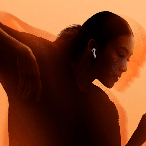 A person with long hair in a ponytail wears AirPods, showcasing an immersive audio experience as they move gracefully against an orange background, with motion blur emphasizing movement and expression.