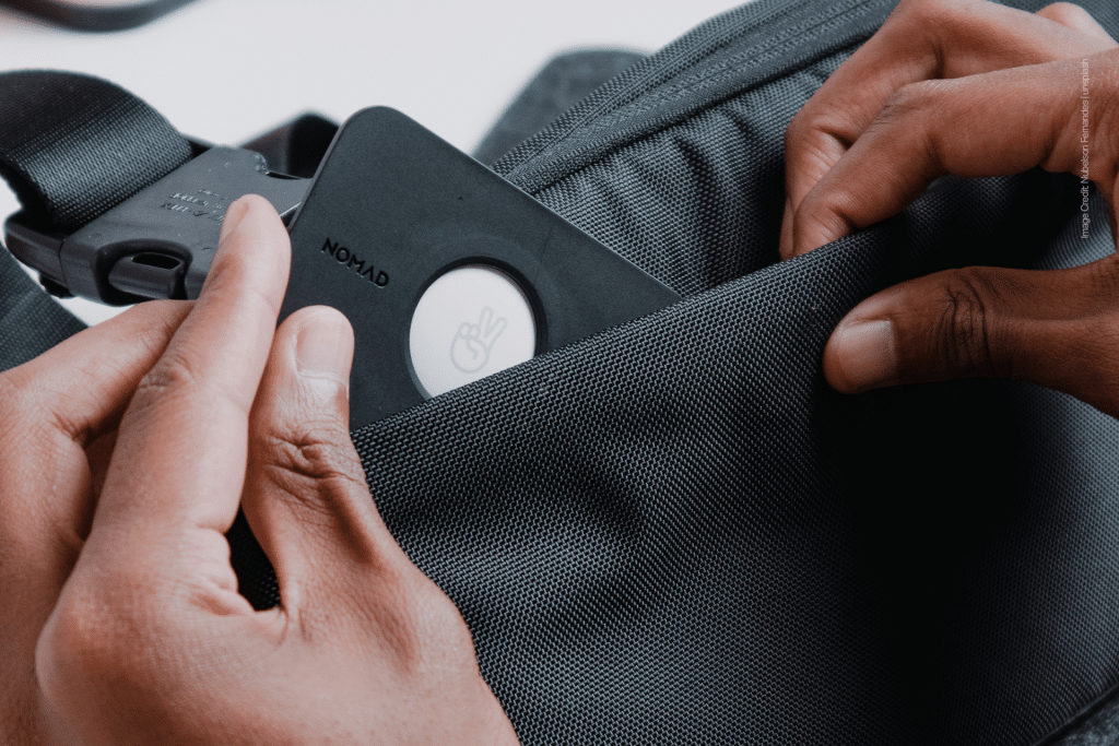 Close-up of hands placing a black device with a peace sign icon, similar to an AirTag, into the front pocket of a gray fabric bag—a smart choice for travelers looking to avoid lost luggage.