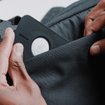 Close-up of hands placing a black device with a peace sign icon, similar to an AirTag, into the front pocket of a gray fabric bag—a smart choice for travelers looking to avoid lost luggage.