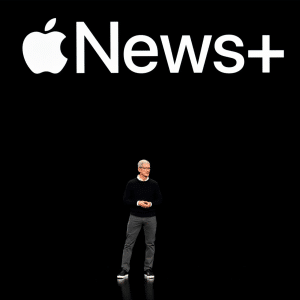 A person stands on a dark stage beneath a large "Apple News+" logo, as the UK launch introduces curated news and immersive Audio stories with the Apple symbol followed by the word "News+" in white text.