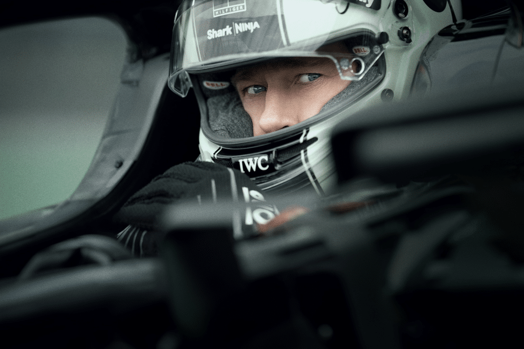 A race car driver wearing a helmet and gloves sits in the cockpit, looking intently ahead. The focus is on the driver's eyes, with car details and helmet logos visible—capturing the intensity of Apple’s $300 Million F1 movie.