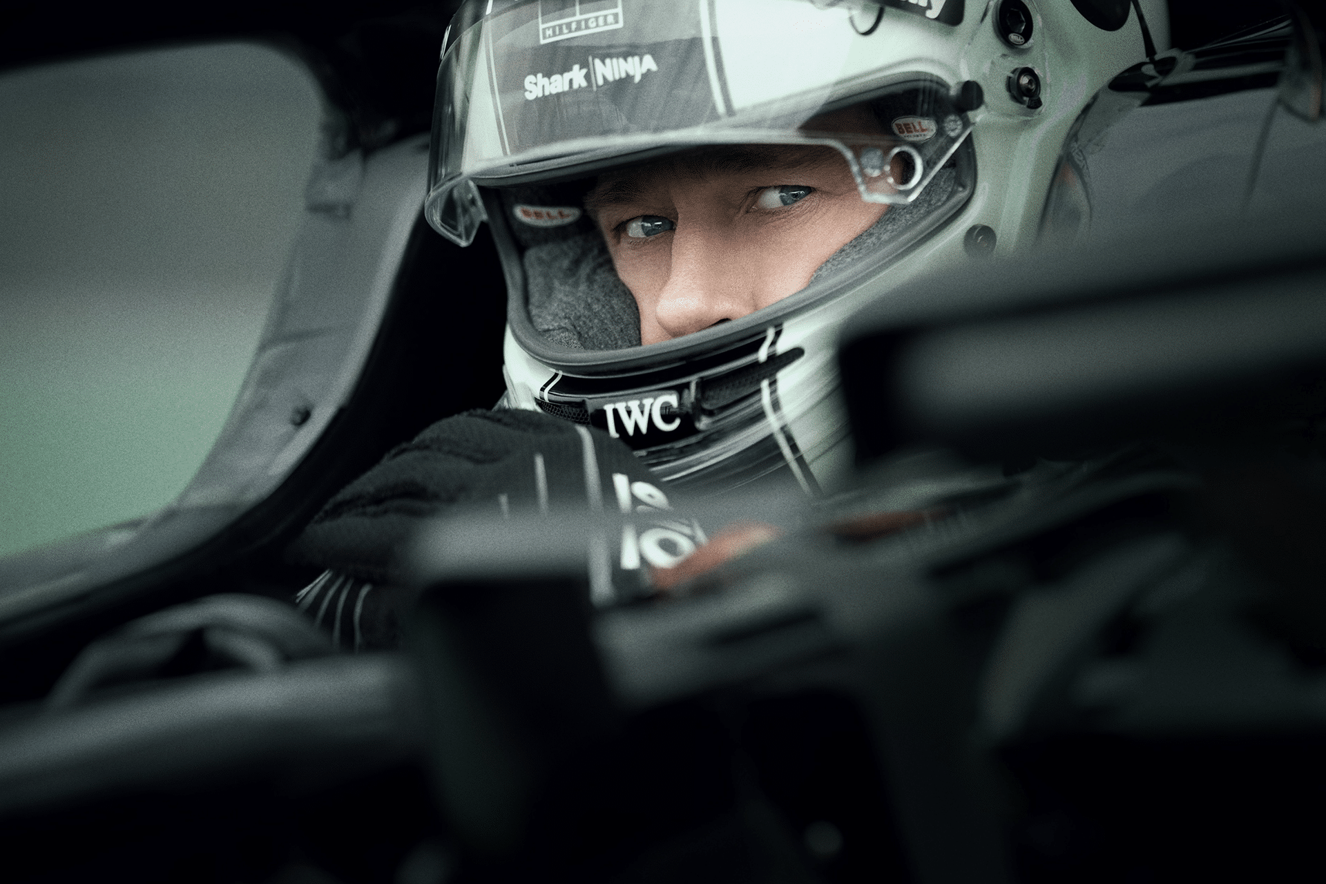 A race car driver wearing a helmet and gloves sits in the cockpit, looking intently ahead. The focus is on the driver's eyes, with car details and helmet logos visible—capturing the intensity of Apple’s $300 Million F1 movie.