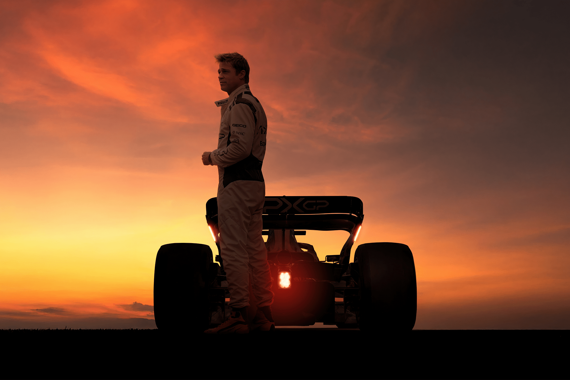 A racecar driver in a racing suit stands next to a formula car at sunset, the dramatic orange sky hinting at scenes from the upcoming Apple F1 Movie, with the car’s taillights illuminated.
