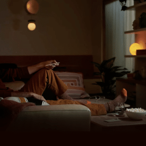 A man wearing headphones relaxes on a sofa, enjoying a Home Theater experience in a cozy, dimly lit living room. He holds a remote while watching TV, with popcorn and a drink on the table in front of him.
