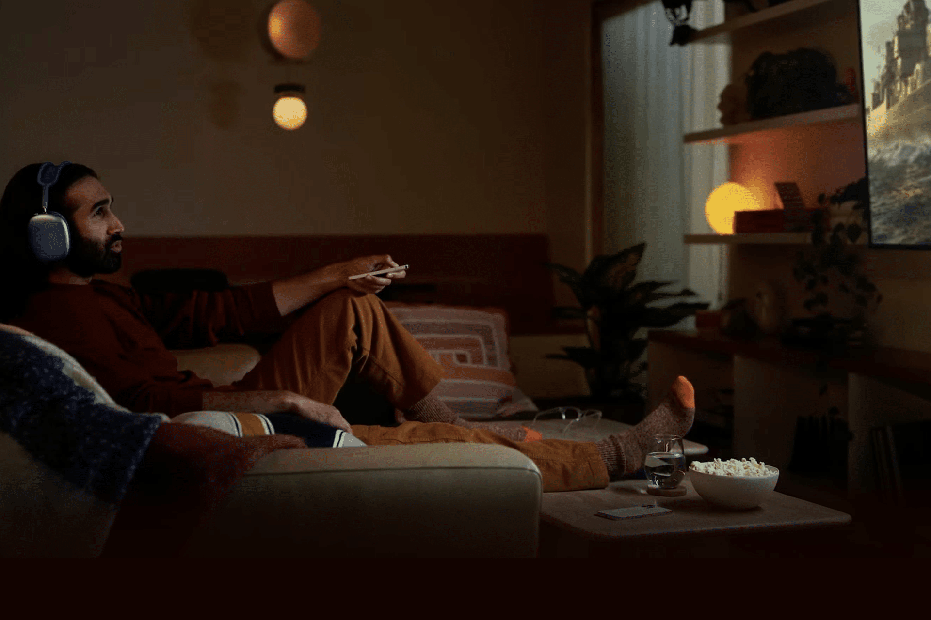 A man wearing headphones relaxes on a sofa, enjoying a Home Theater experience in a cozy, dimly lit living room. He holds a remote while watching TV, with popcorn and a drink on the table in front of him.