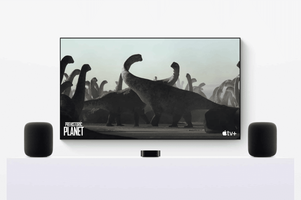 A TV screen with tvOS 26 displays a "Prehistoric Planet" scene on Apple TV+, showing large dinosaurs. Two AirPlay Speakers flank the TV, all set on a white surface—perfect for an immersive home theater experience.