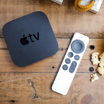 An Apple TV device running tvOS 26 and its remote sit on a wooden table next to a yellow bowl of popcorn, with scattered pieces nearby—setting the scene for the perfect Home Theater night.