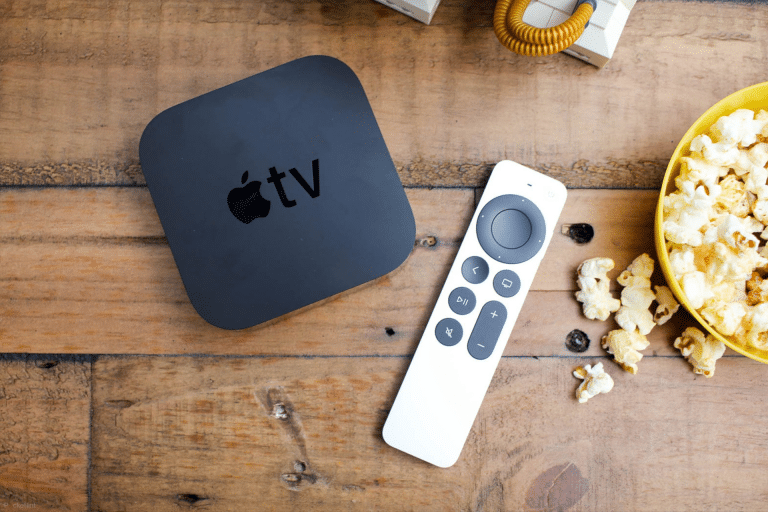 An Apple TV device running tvOS 26 and its remote sit on a wooden table next to a yellow bowl of popcorn, with scattered pieces nearby—setting the scene for the perfect Home Theater night.