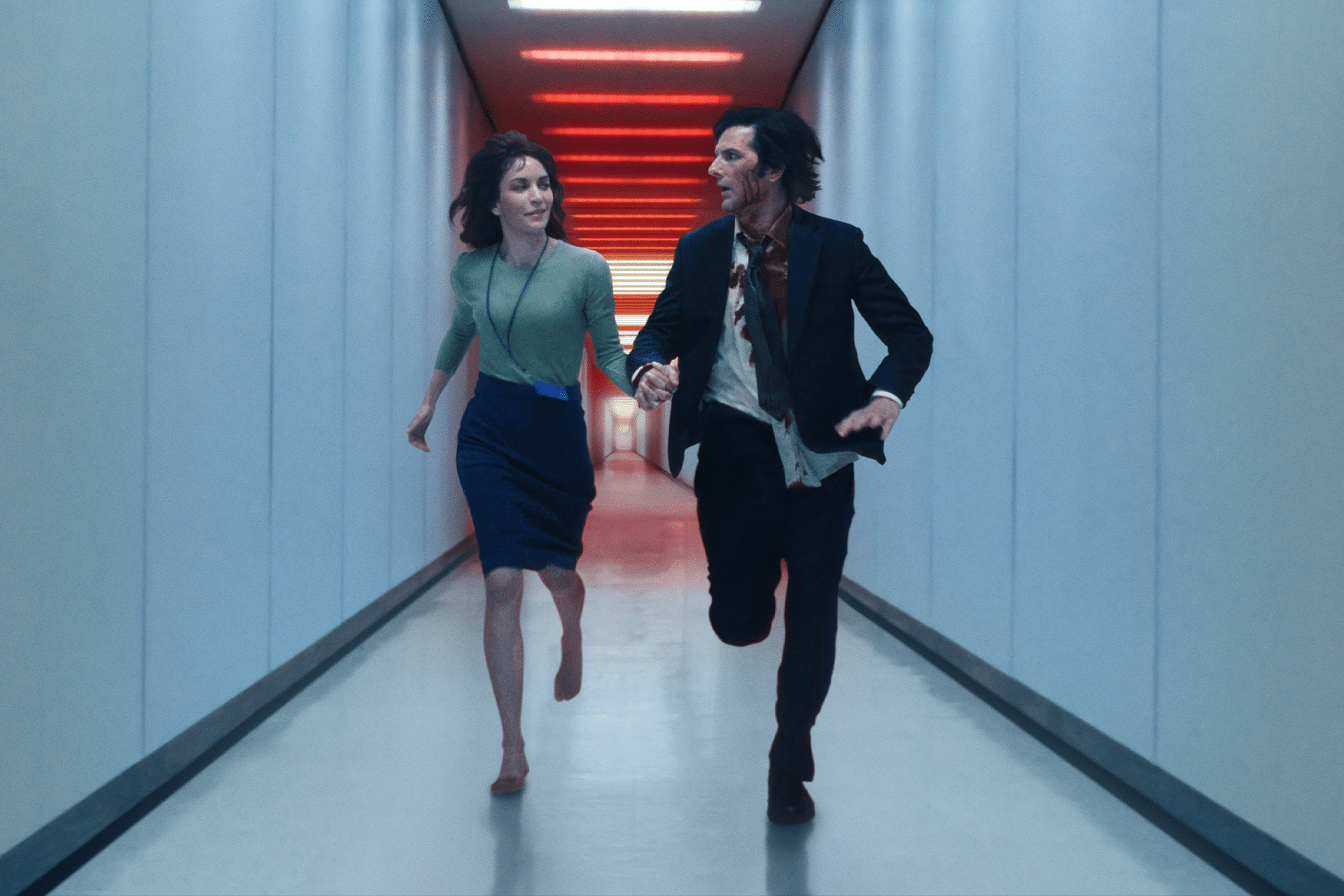 A woman and a man run side by side down a brightly lit, futuristic hallway with red ceiling lights, reminiscent of Severance, looking at each other as they move forward.