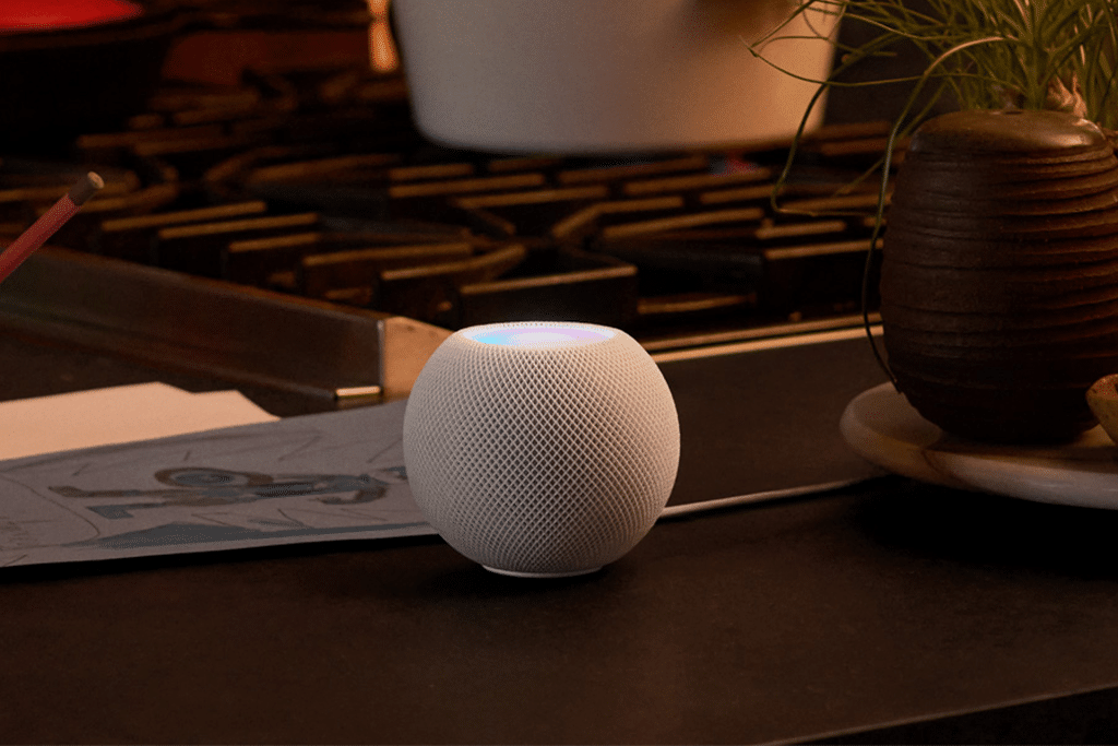 A white Apple smart speaker, possibly the HomePod Mini 2 (2025), with a glowing top sits on a kitchen counter near a stove, beside a potted plant and some papers.