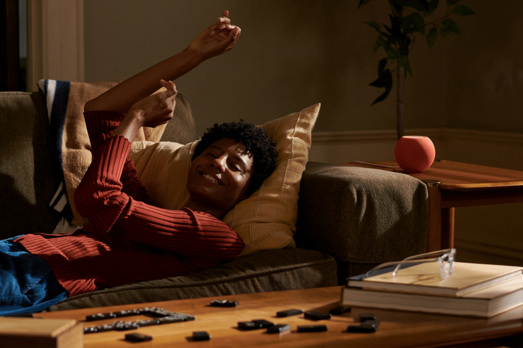 A person smiles and stretches while reclining on a couch, resting their head on a pillow. On the coffee table are books, glasses, dominoes, a remote control, and an Apple smart speaker—possibly the HomePod Mini 2—adding modern flair for 2025.