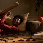 A person smiles and stretches while reclining on a couch, resting their head on a pillow. On the coffee table are books, glasses, dominoes, a remote control, and an Apple smart speaker—possibly the HomePod Mini 2—adding modern flair for 2025.