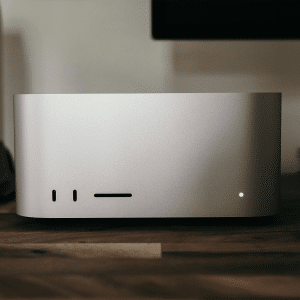 A sleek silver Mac Studio sits on a wooden desk, designed to streamline desktop productivity. With minimal ports and a single small light on the front, it’s surrounded by a blurred monitor and other tech accessories in the background.