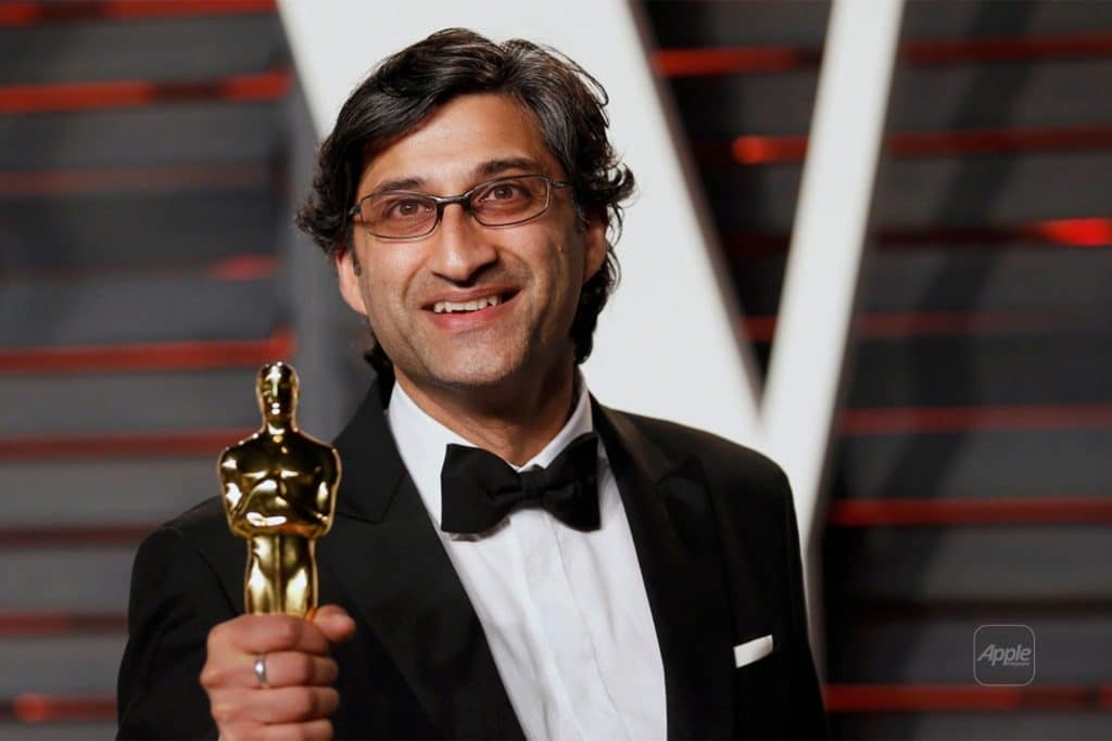 A man in a tuxedo and glasses smiles while holding an Oscar statuette in his right hand at an awards event, reminiscent of Asif Kapadia, renowned for blending fact with fiction in documentary filmmaking.