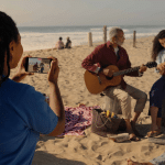 A person captures immersive memories with a phone, taking a spatial photo of two people playing guitar and ukulele on a sandy beach near the ocean, surrounded by blankets and a picnic basket.
