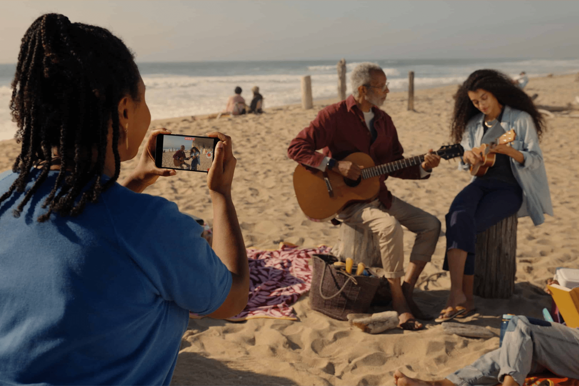 A person captures immersive memories with a phone, taking a spatial photo of two people playing guitar and ukulele on a sandy beach near the ocean, surrounded by blankets and a picnic basket.