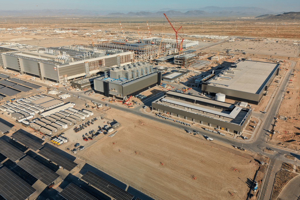 Aerial view of a large industrial construction site in a desert area, showing several partially built buildings, cranes, construction equipment, vehicles, and solar panels in the foreground at TSMC’s Arizona plants for advanced chip production. Mountains are visible in the background.
