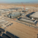 Aerial view of a large industrial construction site in a desert area, showing several partially built buildings, cranes, construction equipment, vehicles, and solar panels in the foreground at TSMC’s Arizona plants for advanced chip production. Mountains are visible in the background.