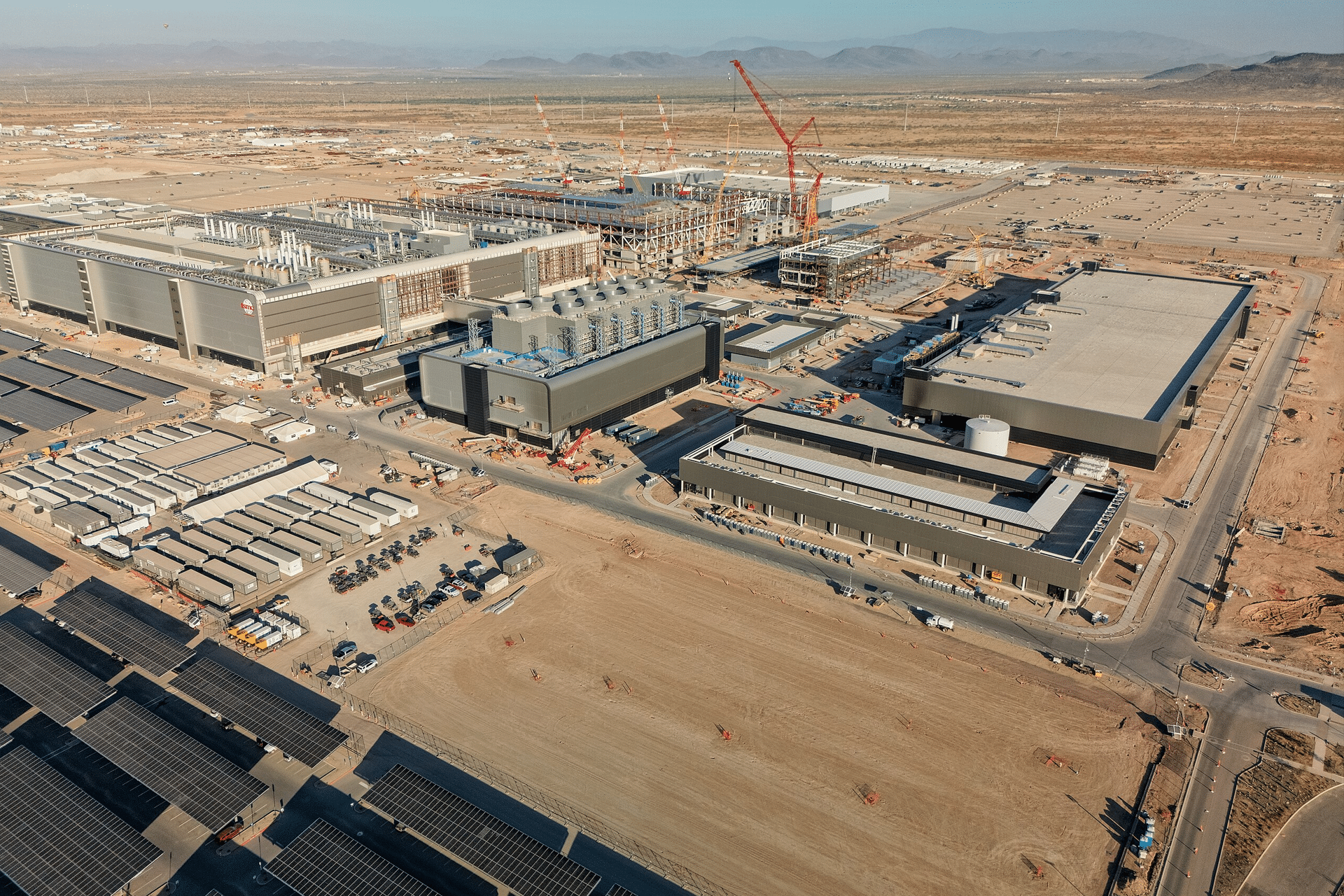 Aerial view of a large industrial construction site in a desert area, showing several partially built buildings, cranes, construction equipment, vehicles, and solar panels in the foreground at TSMC’s Arizona plants for advanced chip production. Mountains are visible in the background.