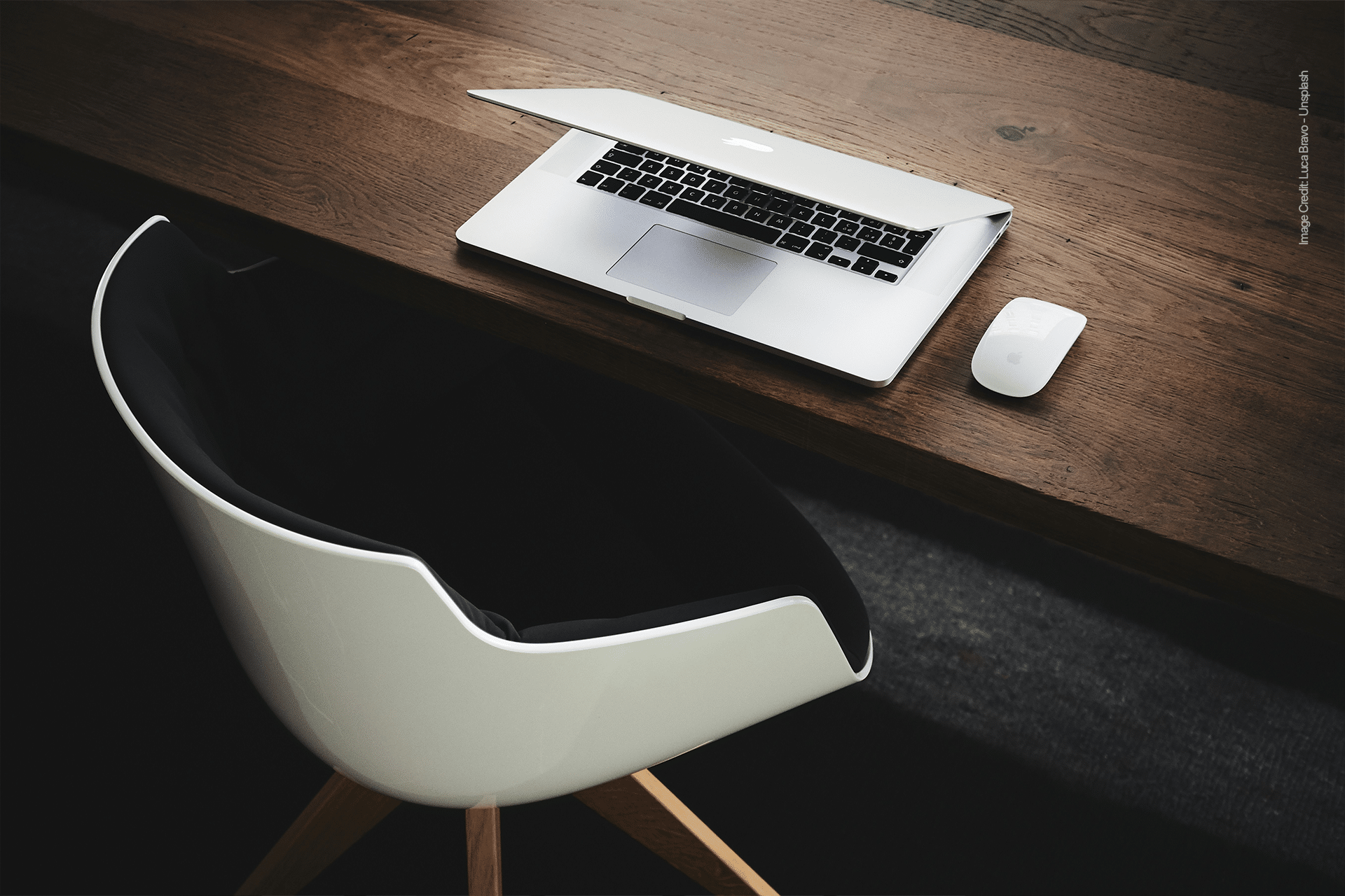 A white modern chair sits in front of a wooden desk with a partially closed laptop and a white wireless mouse, creating a minimal, clean aesthetic—an ideal spatial workspace for peak focus.