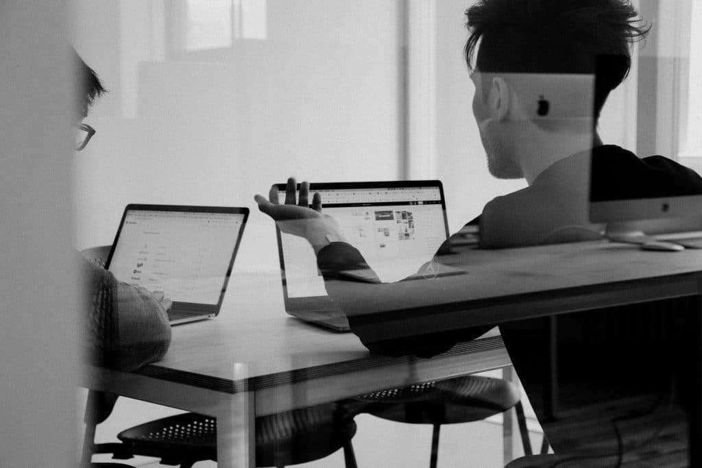 Two people sit at a table with open laptops, discussing work. One gestures toward their screen, both immersed in peak focus. The scene is reflected in glass, adding a layered effect to the modern spatial workspace.