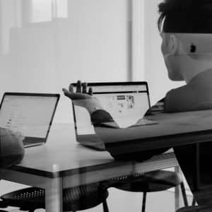 Two people sit at a table with open laptops, discussing work. One gestures toward their screen, both immersed in peak focus. The scene is reflected in glass, adding a layered effect to the modern spatial workspace.