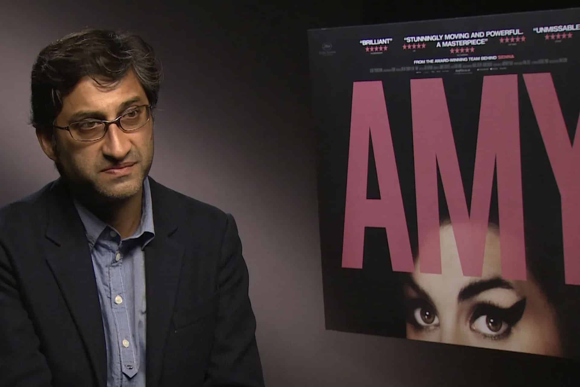 A man in glasses and a dark blazer sits for an interview next to a large poster for Asif Kapadia’s documentary "AMY," featuring a close-up of a woman's eyes and bold pink letters.