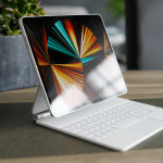 A tablet with a colorful abstract display is attached to a white keyboard and trackpad, positioned on a table beside a small pot of succulents.