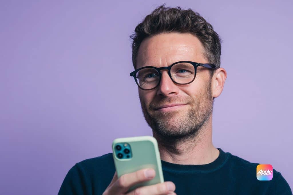 A man with glasses and a beard smiles while holding a smartphone against a purple background. An Apple logo is displayed in the lower right corner of the image.