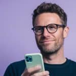 A man with glasses and a beard smiles while holding a smartphone against a purple background. An Apple logo is displayed in the lower right corner of the image.