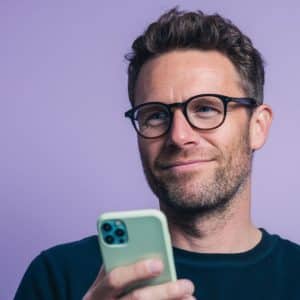 A man with glasses and a beard smiles while holding a smartphone against a purple background. An Apple logo is displayed in the lower right corner of the image.