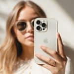 A woman wearing sunglasses holds up a white iPhone, showing the back with triple cameras. She stands in soft natural light, and an Apple logo is visible in the lower right corner of the image.