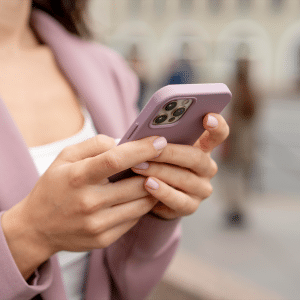 A woman wearing a mauve blazer holds and uses an Apple smartphone with a matching mauve case while standing outdoors on a city street. Her face is partially out of frame, reflecting a human-centered scene with blurred people in the background.