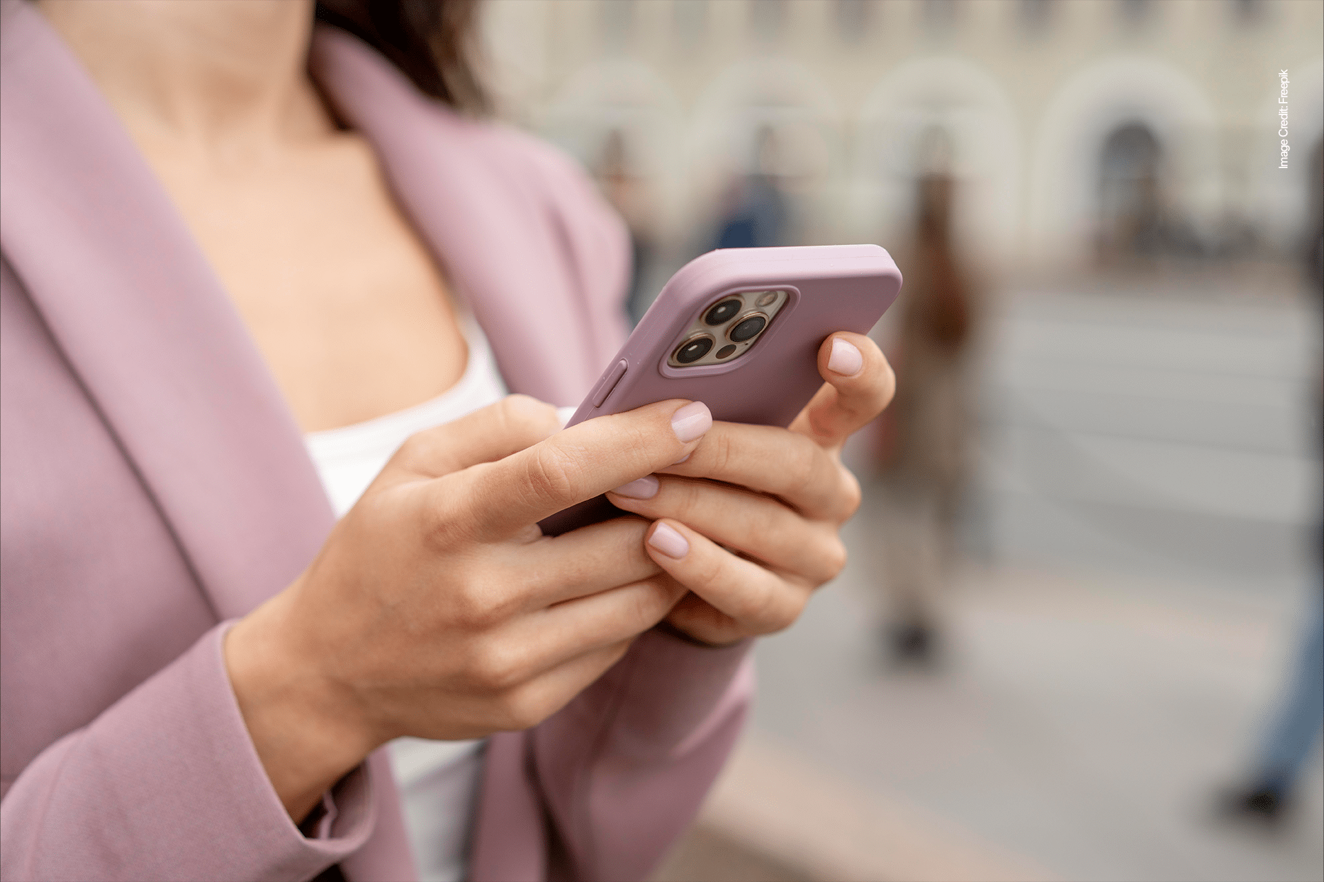 A woman wearing a mauve blazer holds and uses an Apple smartphone with a matching mauve case while standing outdoors on a city street. Her face is partially out of frame, reflecting a human-centered scene with blurred people in the background.