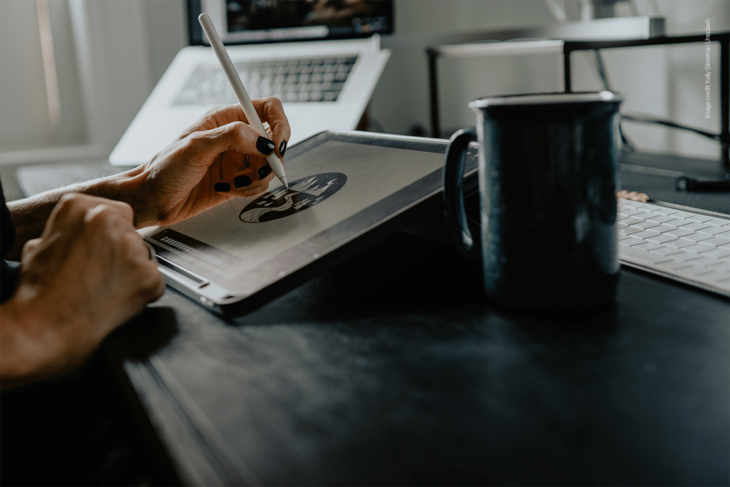 A person uses a stylus to draw on a tablet at a desk, with a mug, keyboard, and laptop nearby in a softly lit workspace—perfect for managing iPad storage or connecting iPad external drives for added convenience.