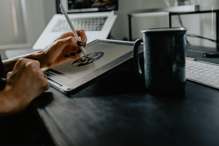 A person uses a stylus to draw on a tablet at a desk, with a mug, keyboard, and laptop nearby in a softly lit workspace—perfect for managing iPad storage or connecting iPad external drives for added convenience.