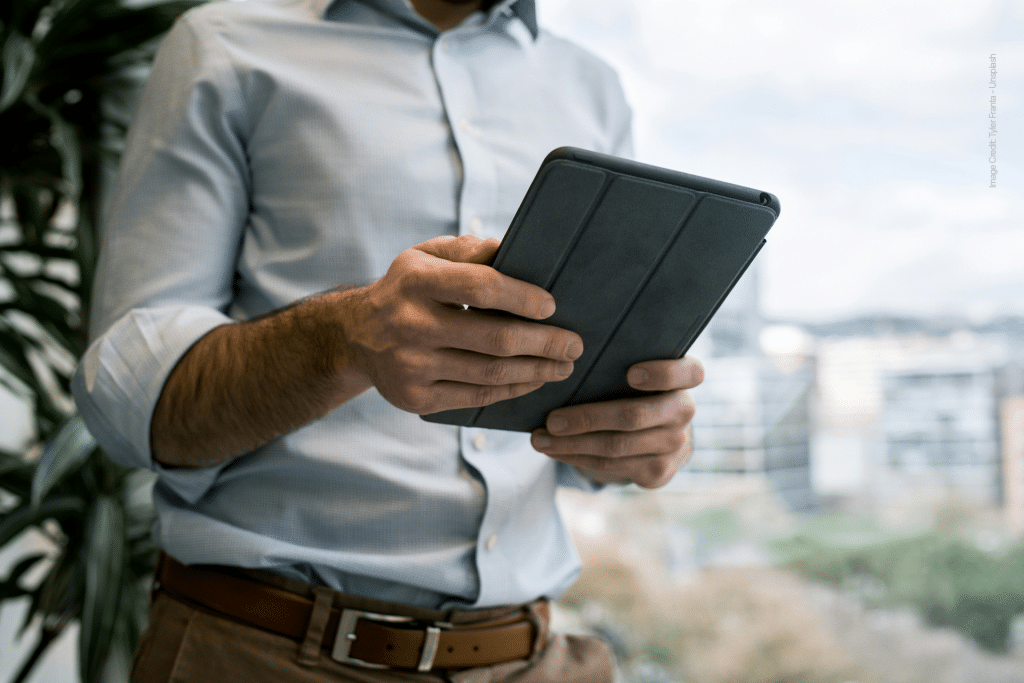 A person in a light blue shirt holds and uses a black tablet indoors, with the iPad home screen and widgets visible, as a blurred cityscape appears through the window in the background.