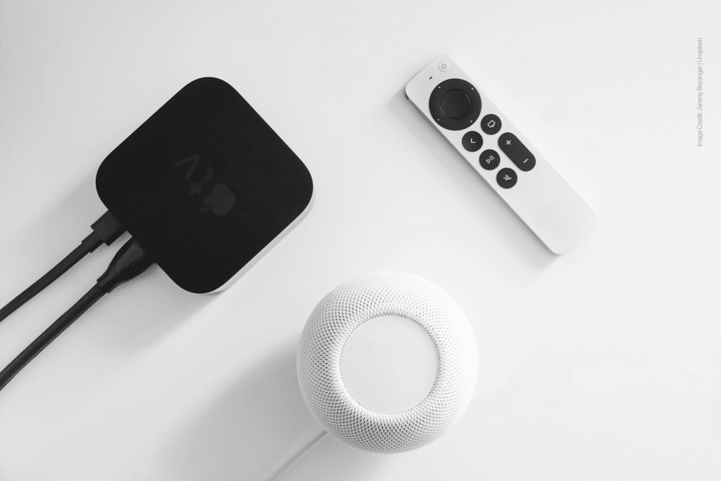 A black Apple TV box running tvOS 18.6 with connected cables, a white Apple TV remote, and a white Apple HomePod mini with HomePod 18.6 on a white surface.