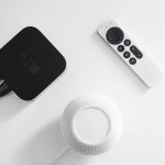 A black Apple TV box running tvOS 18.6 with connected cables, a white Apple TV remote, and a white Apple HomePod mini with HomePod 18.6 on a white surface.