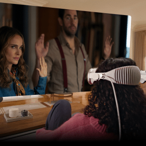 Two people wearing VR headsets sit on a couch in a bright, modern living room, enjoying immersive environments with visionOS as they watch a large virtual screen showing a woman and man raising their hands.