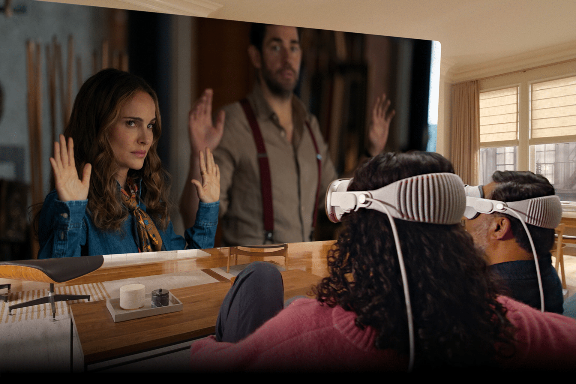 Two people wearing VR headsets sit on a couch in a bright, modern living room, enjoying immersive environments with visionOS as they watch a large virtual screen showing a woman and man raising their hands.