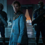 A person in futuristic clothing stands in a dimly lit hall, flanked by two armored guards with large weapons. With a blue tint and an air of intensity, the scene evokes the high-stakes drama found in Apple TV+’s diverse hits.